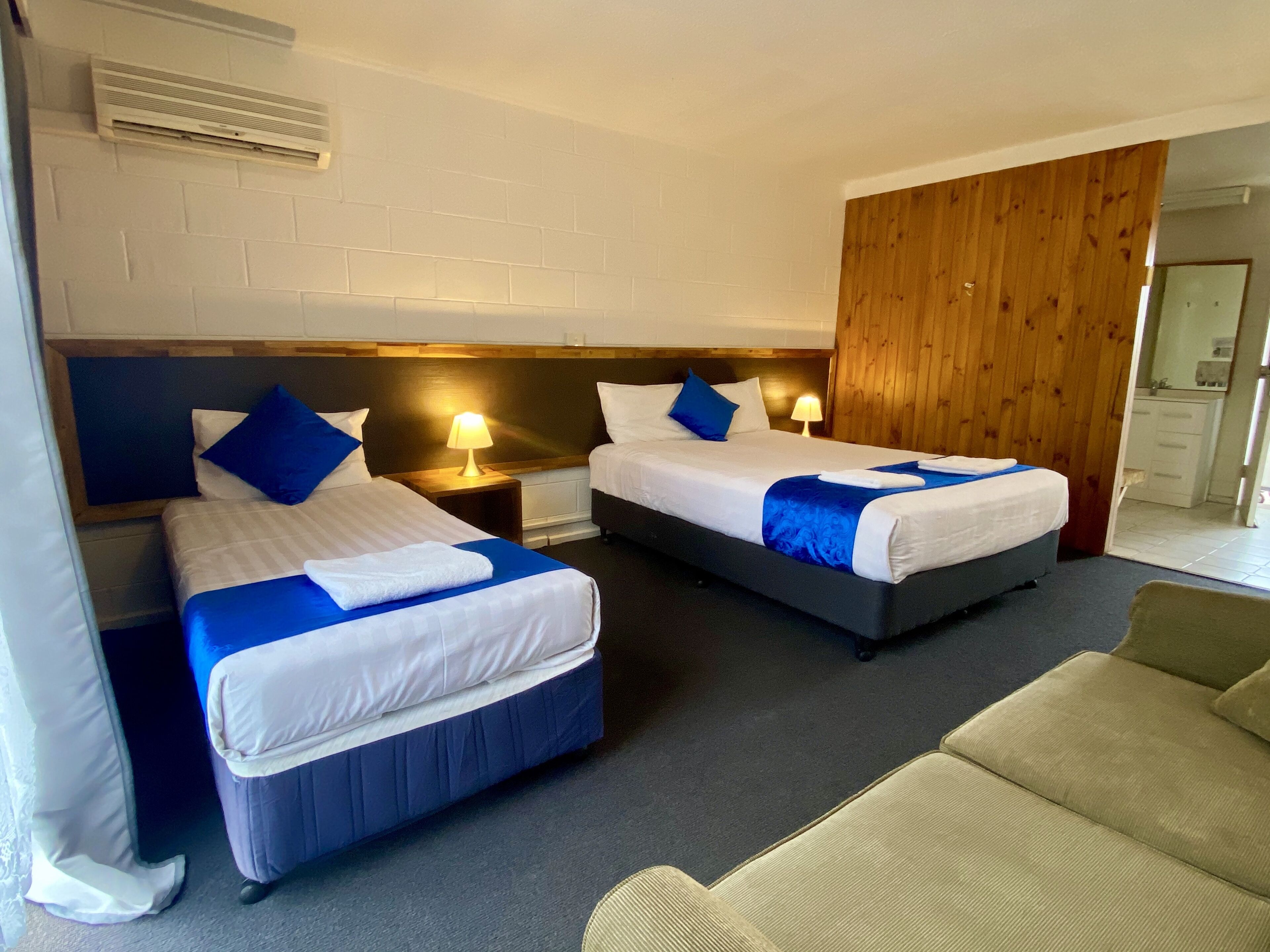 A hotel room with two beds, a wooden headboard, a nightstand with a lamp, and a grey sofa.