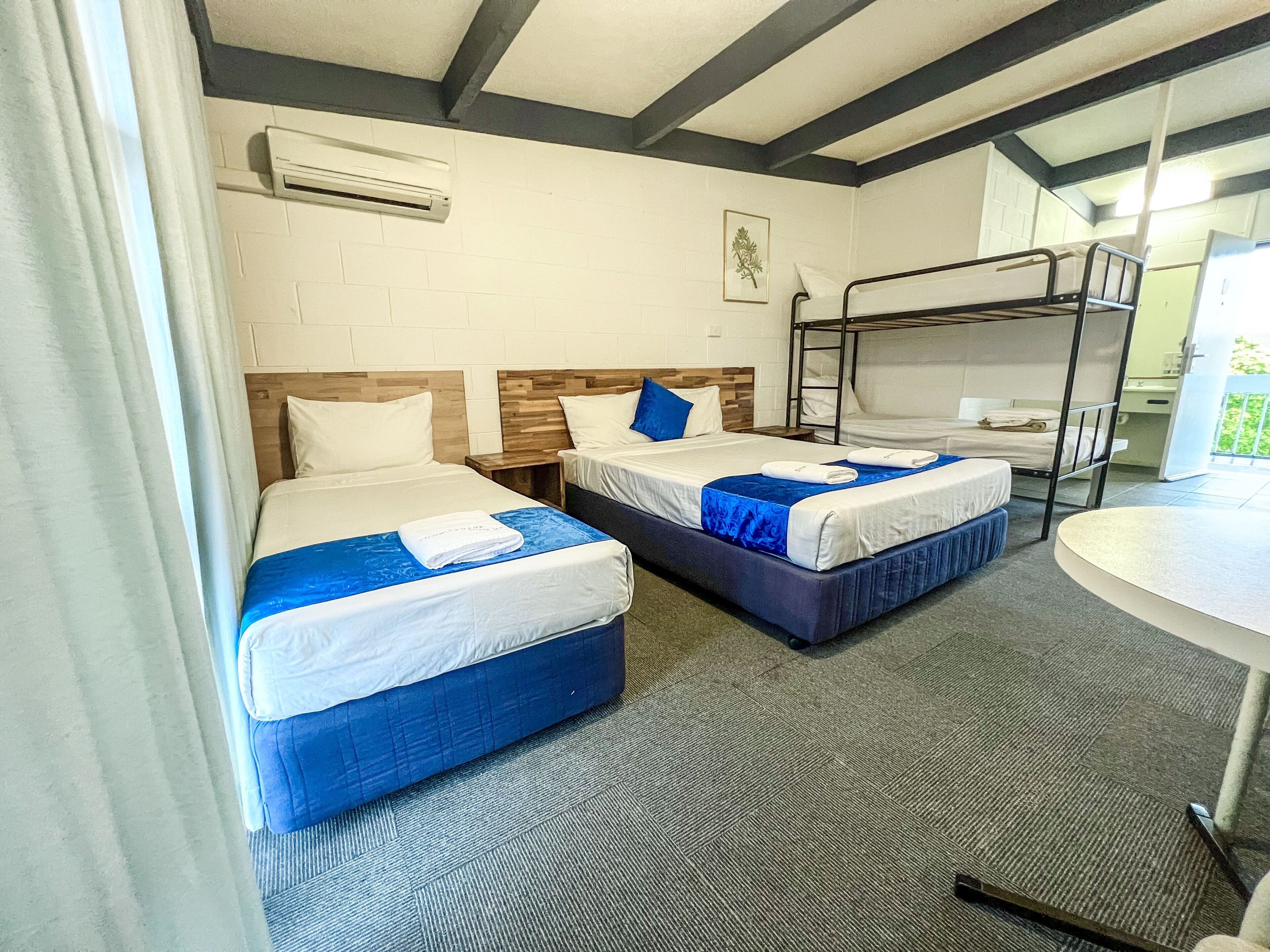 A hotel room with two beds, a bunk bed, a round table, and a window with curtains.
