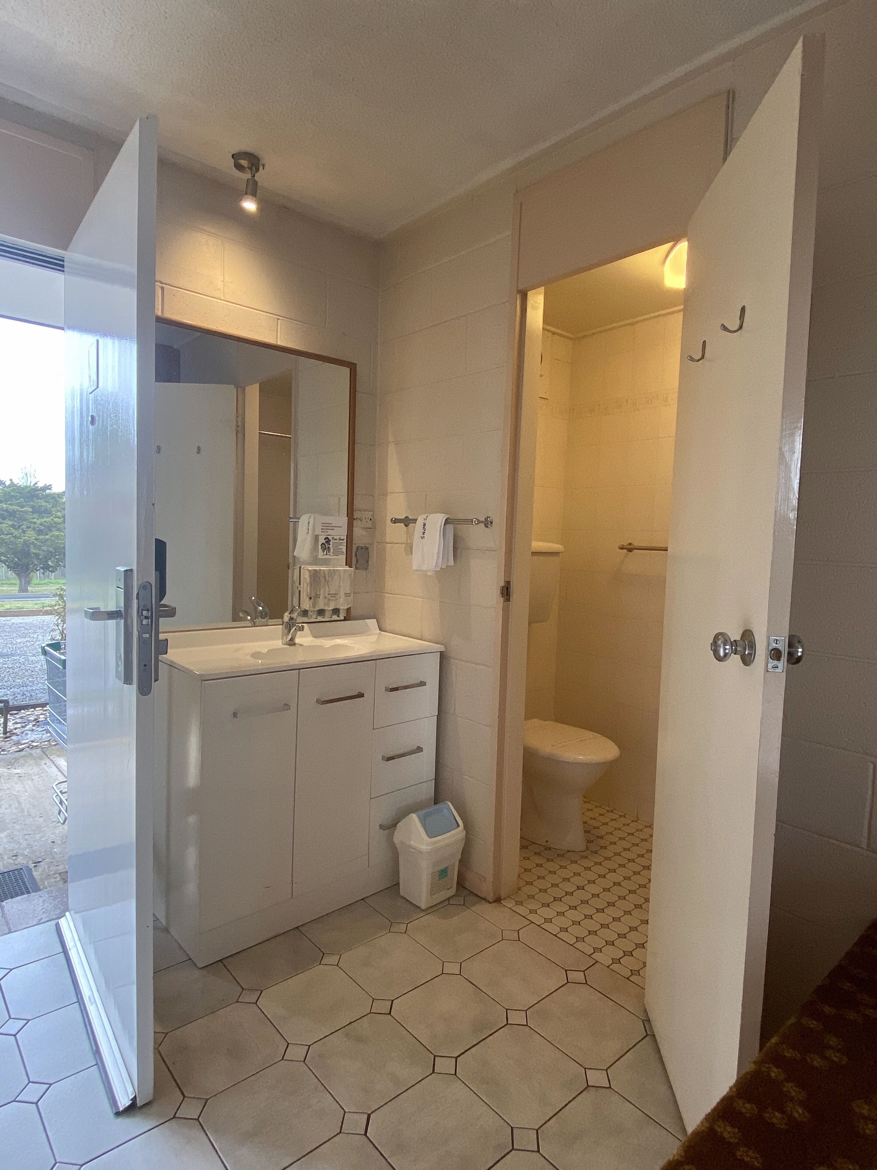 A bathroom with a white sink, mirror, and toilet. There is a trash bin and a towel rack. The floor has hexagonal tiles.