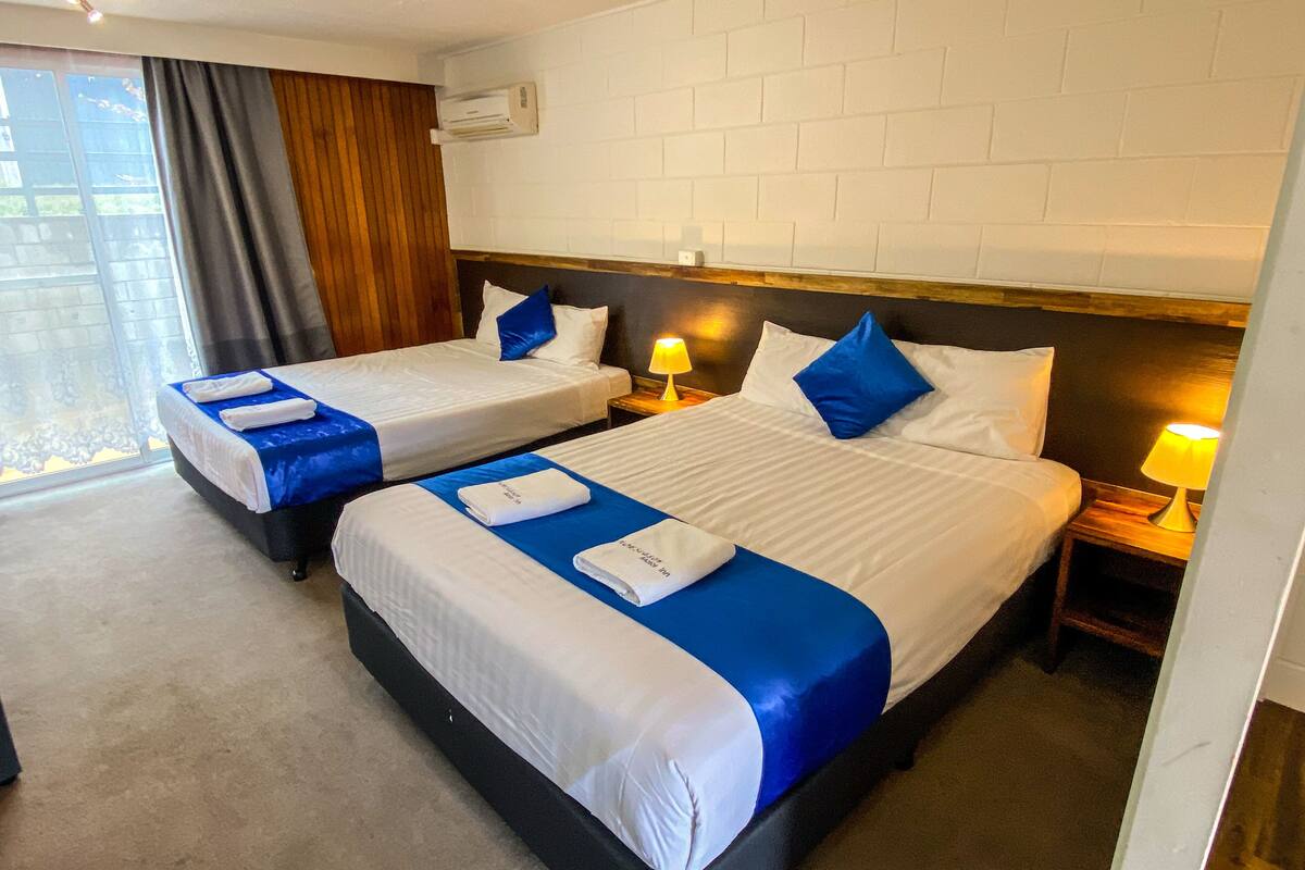A hotel room with two beds, each with white linens and blue accents, a wooden headboard, a window with curtains, and an air conditioning unit on the wall.