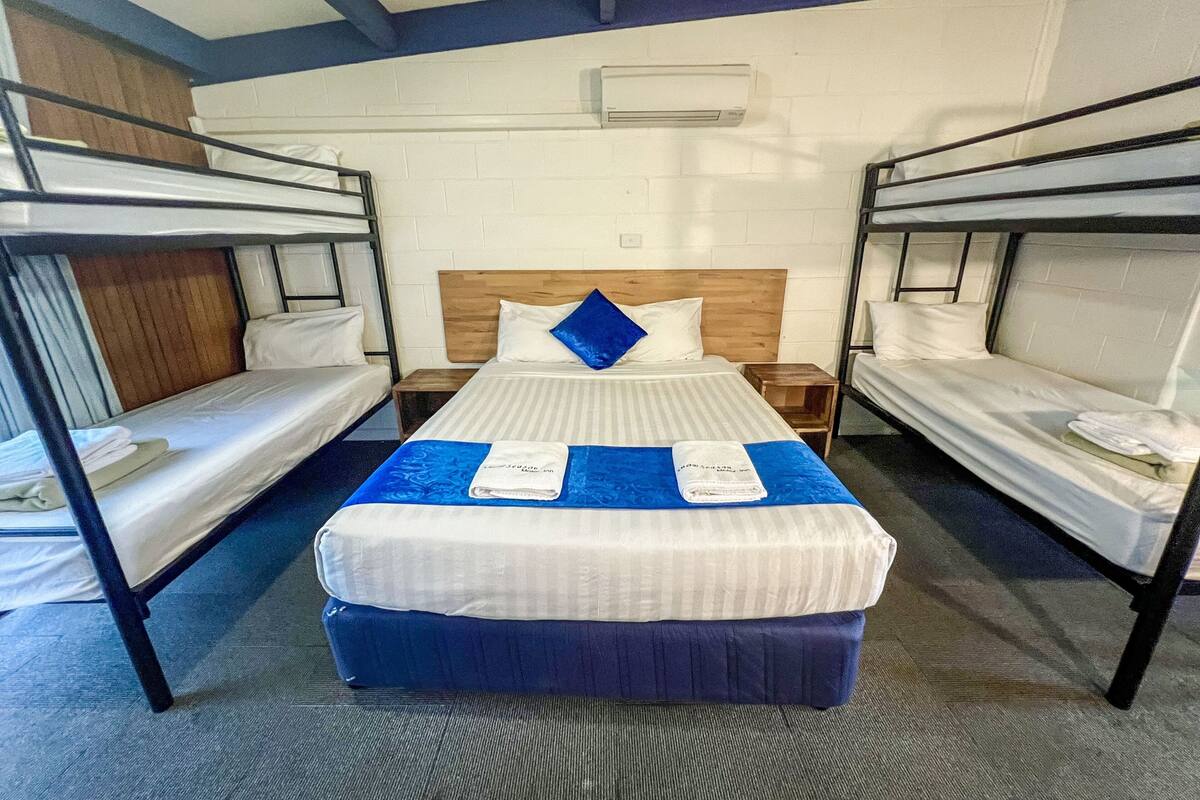 A dormitory room with bunk beds, a wooden headboard, a blue bedspread, and a white wall with an air conditioning unit.
