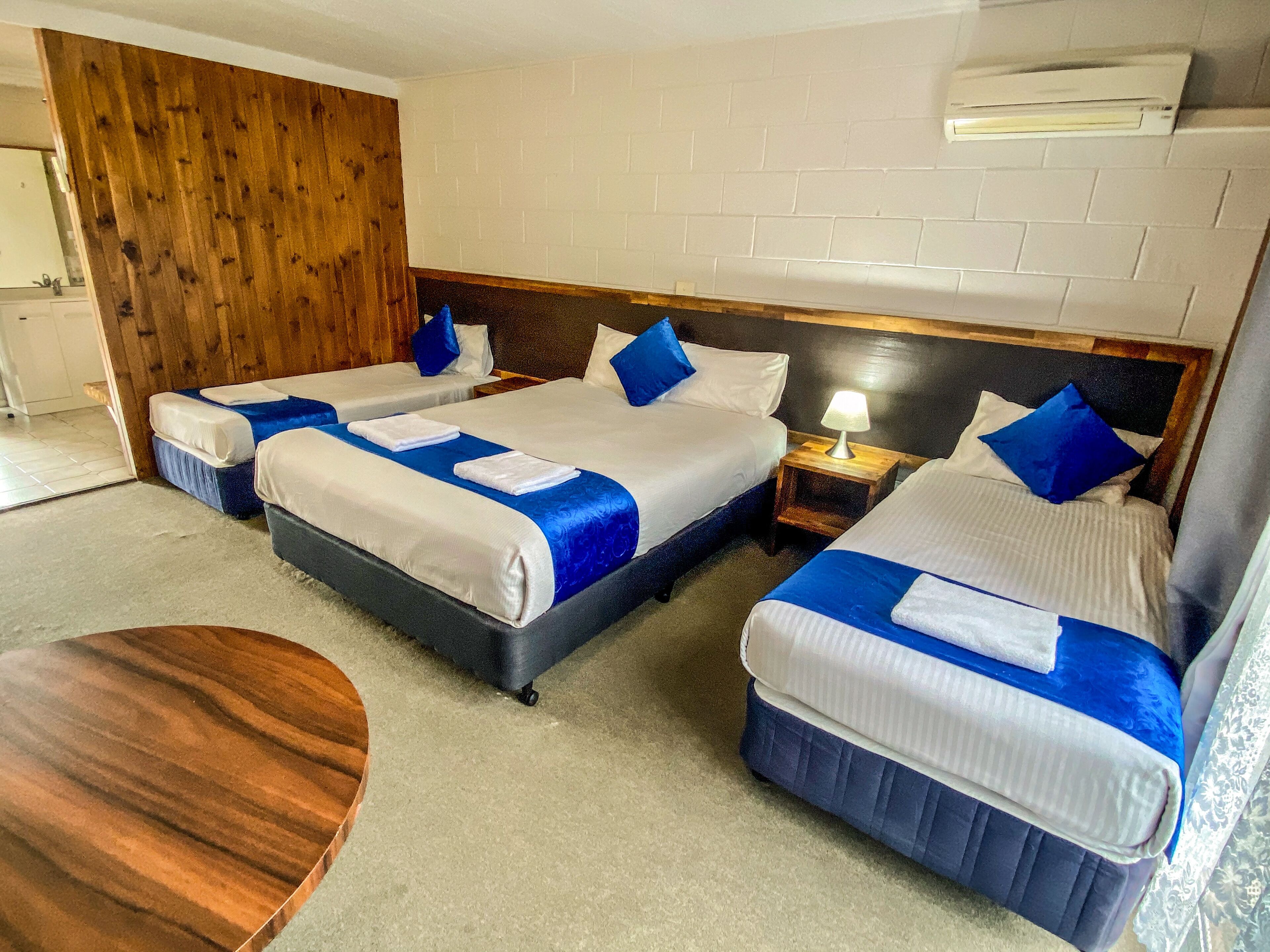 A hotel room with three beds, a wooden wall, a white brick wall, and an air conditioning unit.