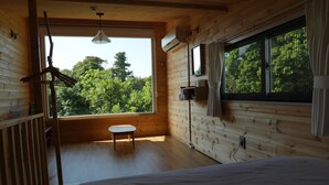 A wooden cabin room with a bed, a small table, and a large window offering a view of trees. - Dabut (Seogwipo)