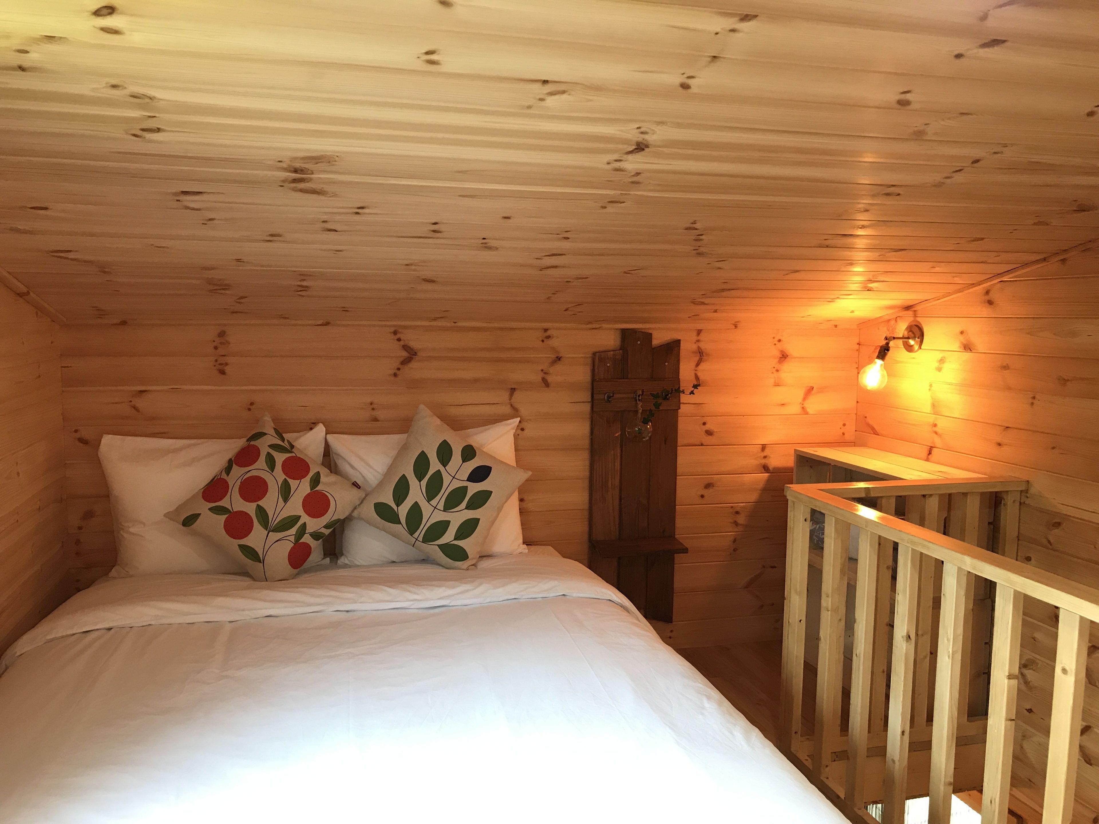 A cozy wooden cabin room with a bed, two pillows with fruit designs, a wooden door, and a small table with a light fixture.