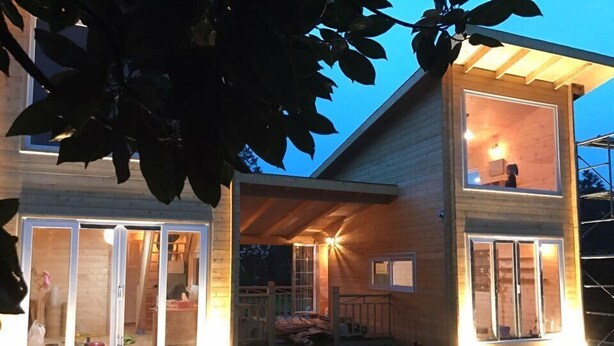 A two-story wooden house with large glass doors and windows, a covered porch, and a well-lit exterior at dusk. - Dabut (Seogwipo)