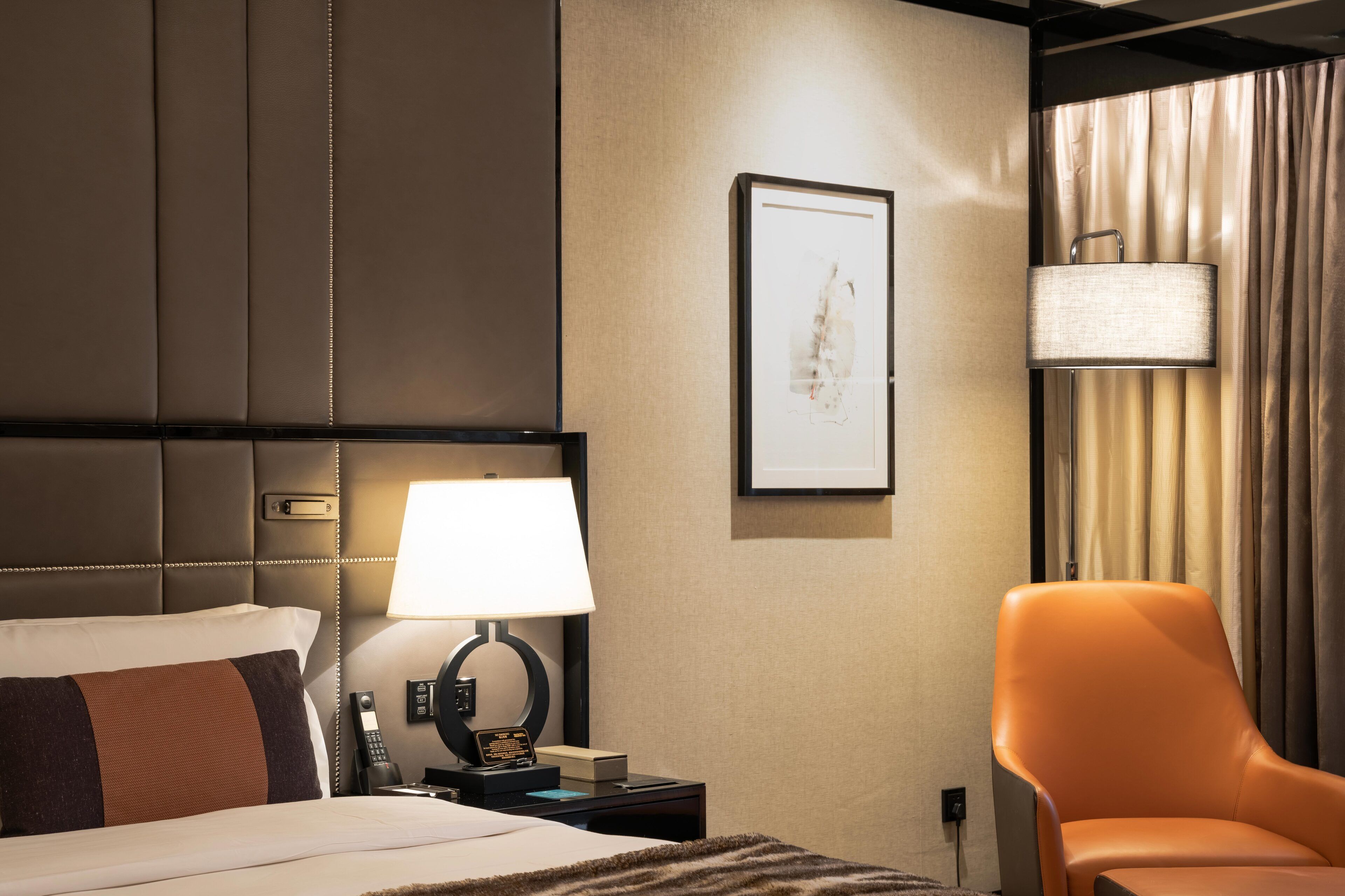 A hotel room with a bed, a nightstand with a lamp, a framed artwork on the wall, and an orange chair.