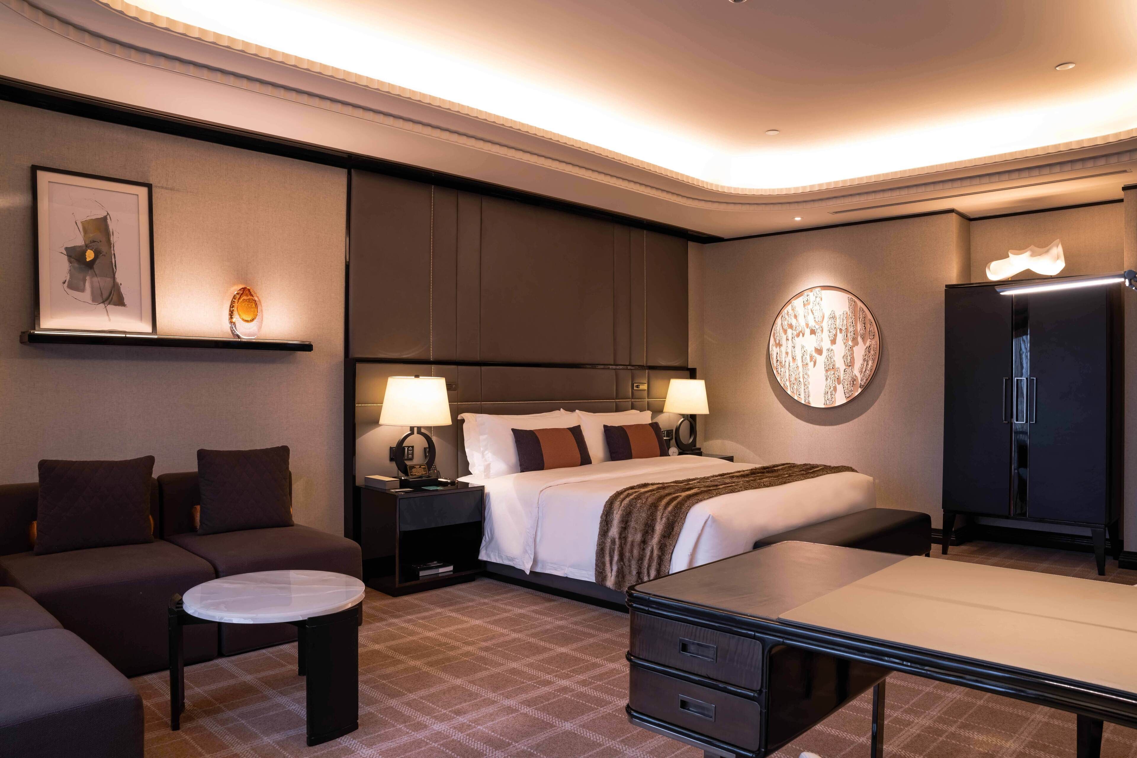 Quarto de hotel moderno com uma cama grande, duas mesas de cabeceira com luminárias, um sofá, uma mesinha redonda e um armário.
