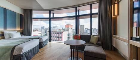 A modern hotel room with two beds, a round table, a sofa, and a large window offering a city view.