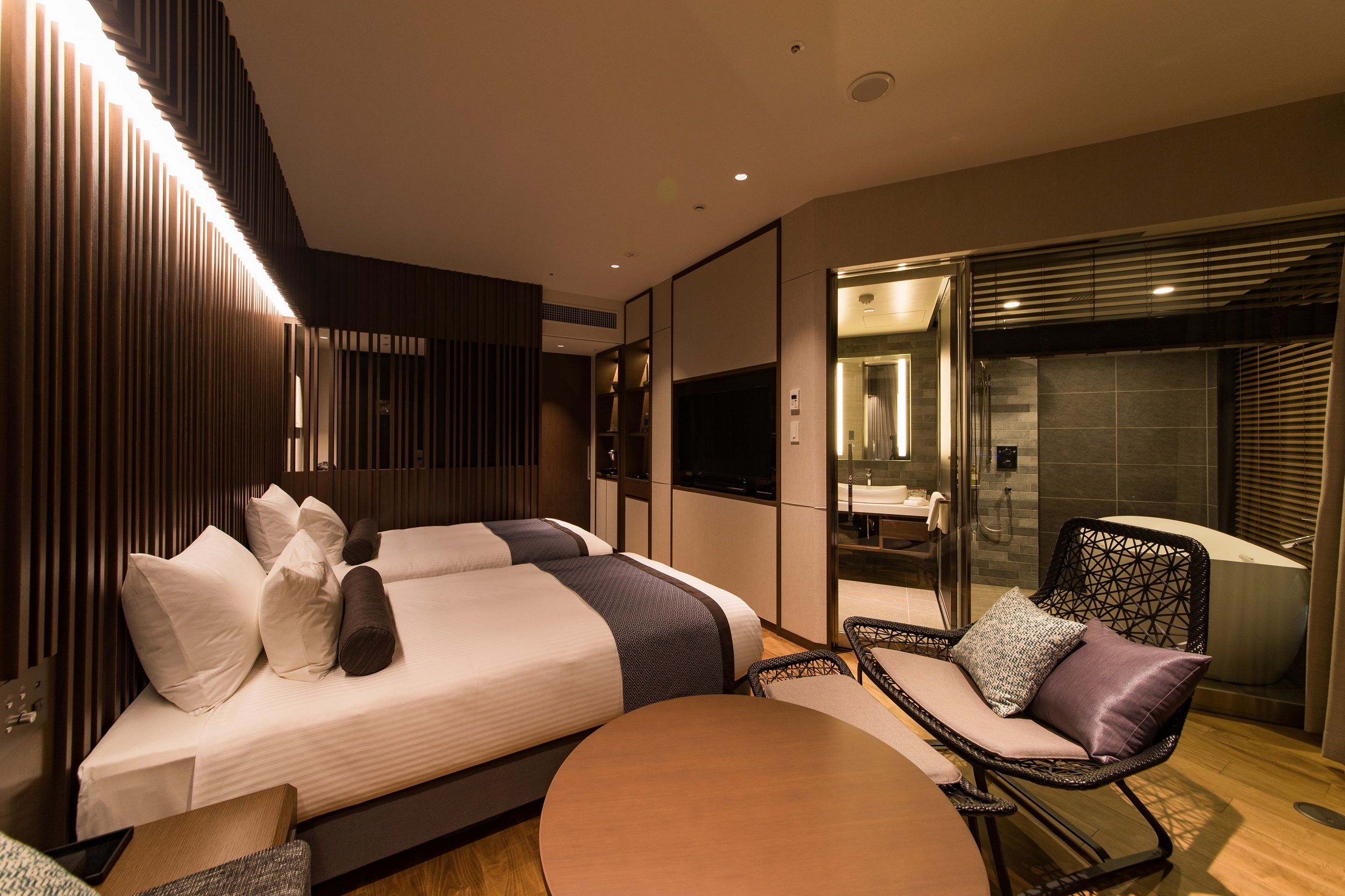A modern hotel room with two beds, a sitting area, and a bathroom with a bathtub.