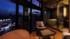 Un intérieur moderne offrant une vue panoramique sur la ville, une salle de bain entièrement vitrée et un coin salon avec une table ronde et deux chaises.