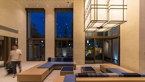 Un espace de détente intérieur moderne, doté de plusieurs canapés et tables, de grandes fenêtres offrant une vue sur la ville et d’un luminaire au design géométrique.