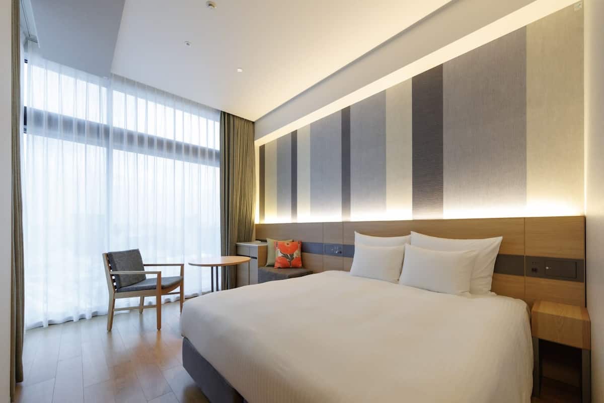 Habitación de hotel moderna con una cama grande, un escritorio, una silla y vistas a la ciudad a través de amplios ventanales.
