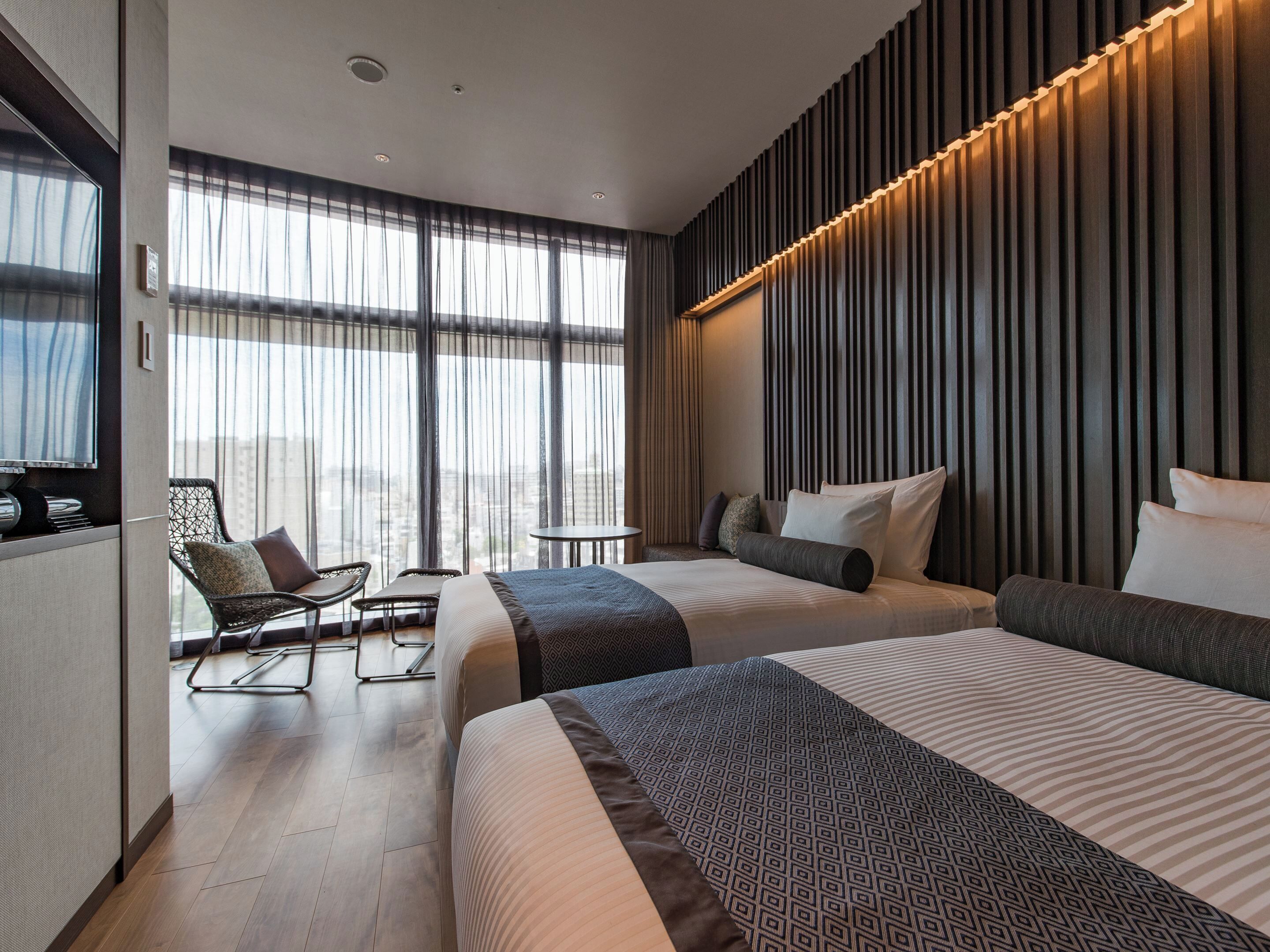 A modern hotel room with two beds, a seating area with chairs, a small table, and large windows with curtains.