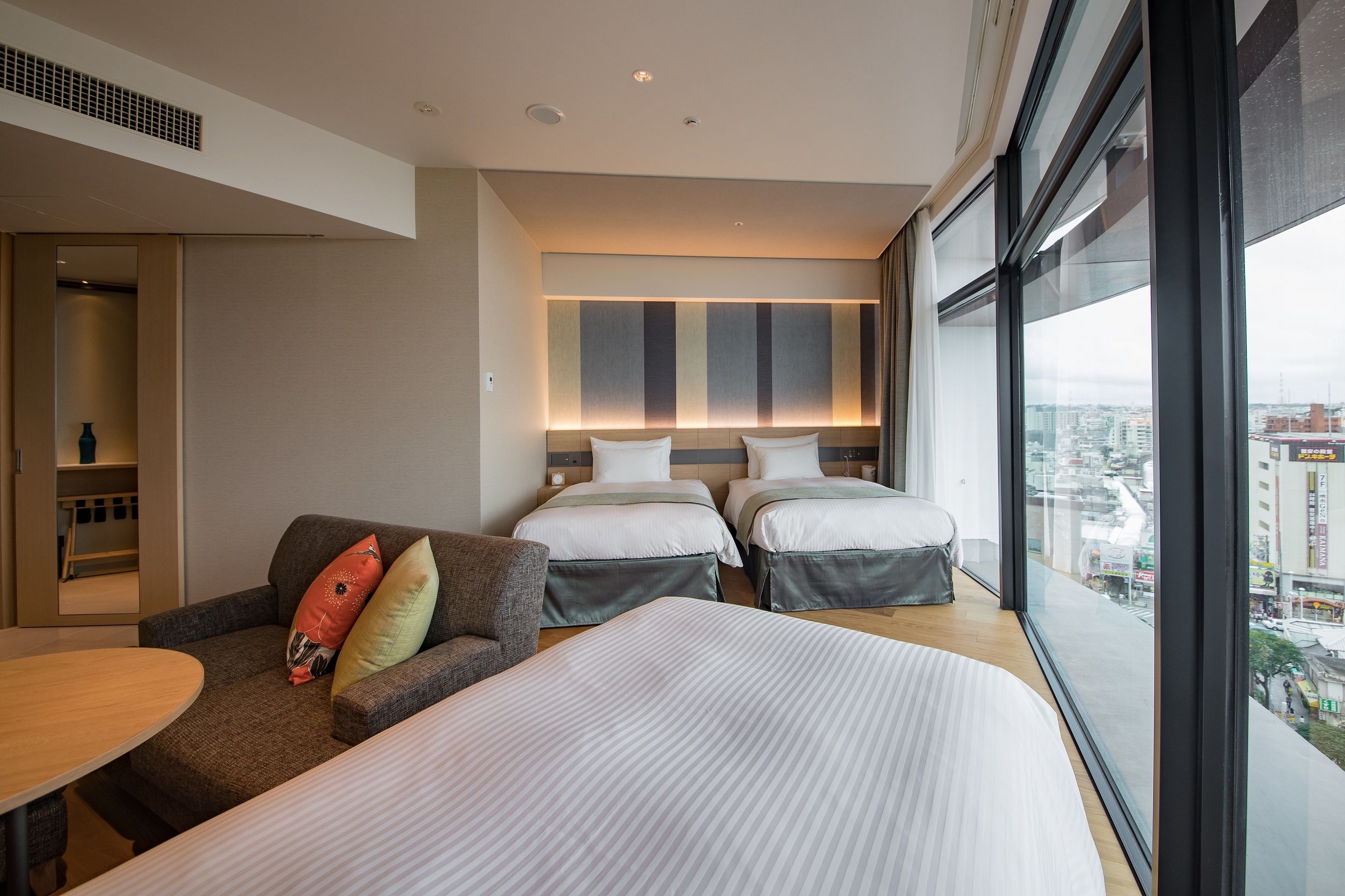 A modern hotel room with a large bed, a sofa, and a small table. There is a city view through the window.