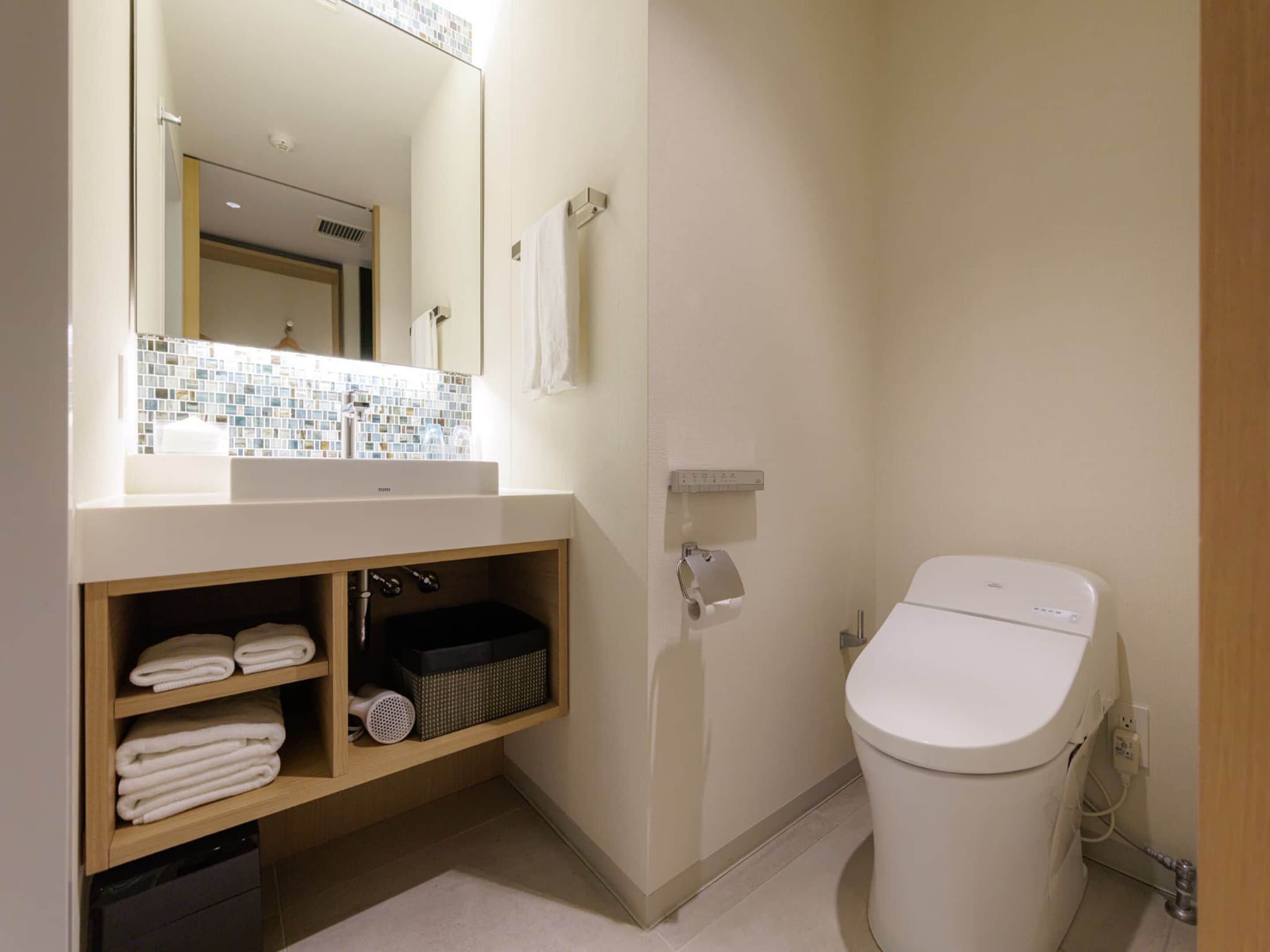 Et moderne badeværelse med hvid vask, spejl og toilet. Der er hylder med håndklæder og en kurv.