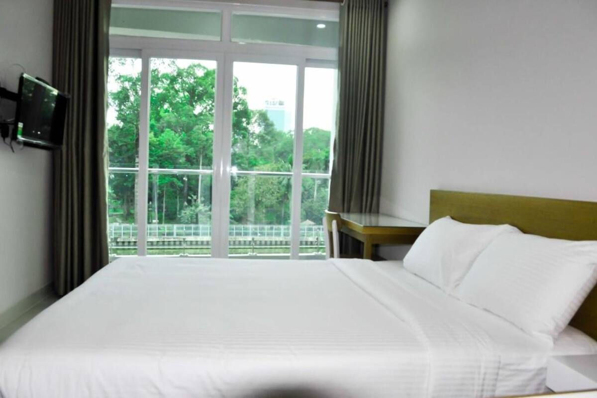 A hotel room with a large bed, a television mounted on the wall, and a view of greenery outside through a large window.