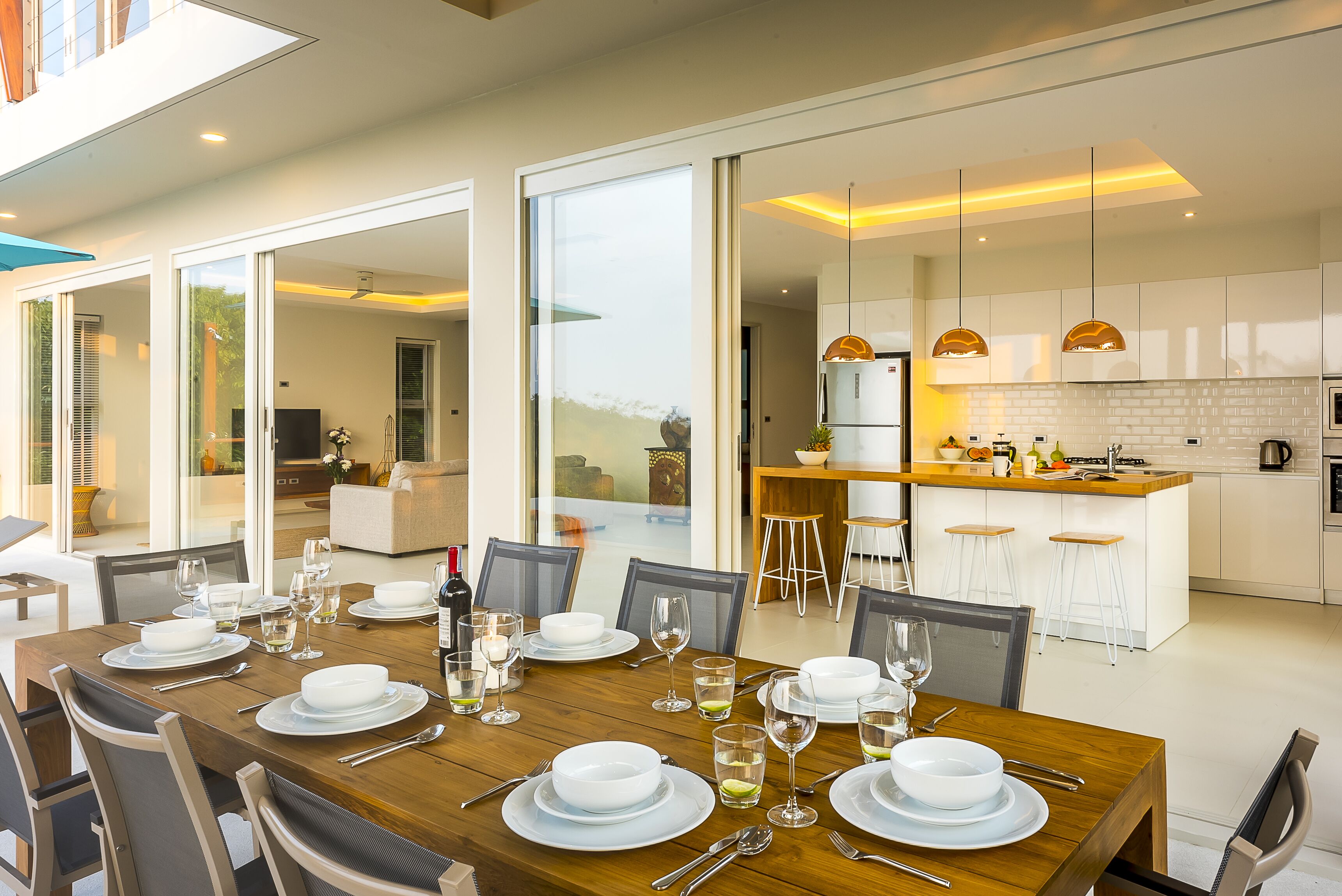 A modern dining area with a wooden table set for a meal, a kitchen with white cabinets, and a large sliding glass door leading to an outdoor area.