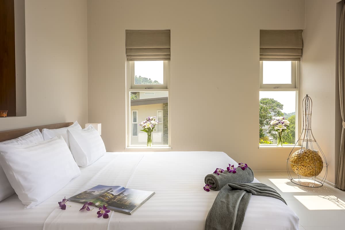 Une chambre d’hôtel avec un grand lit, deux fenêtres avec des rideaux, un luminaire suspendu et une vue sur la verdure.