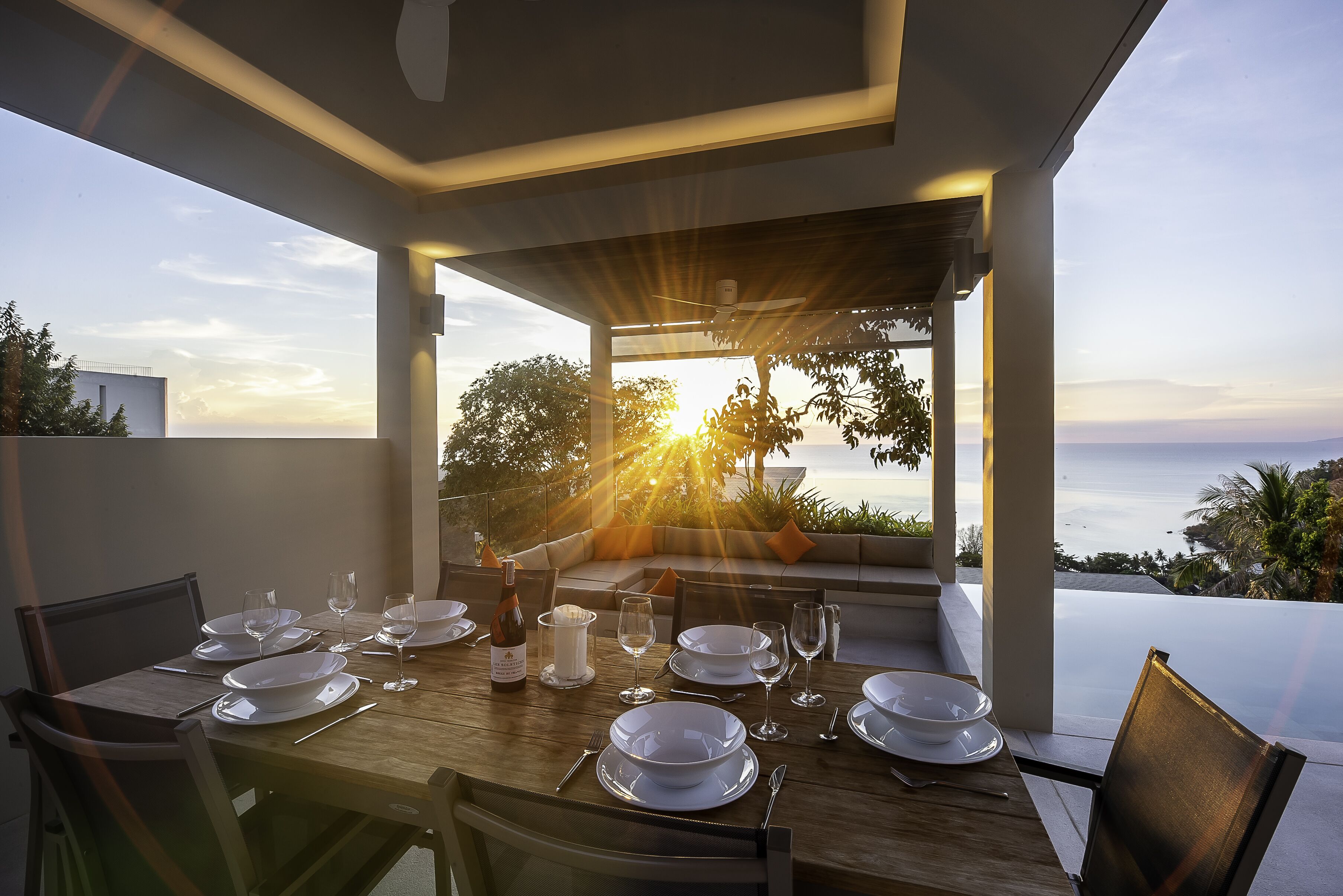 A dining table set for six with white plates, glasses, and cutlery, overlooking a scenic ocean view.