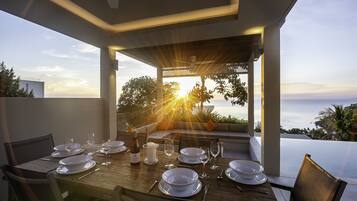 A dining table set for six with white plates, glasses, and cutlery, overlooking a scenic ocean view.