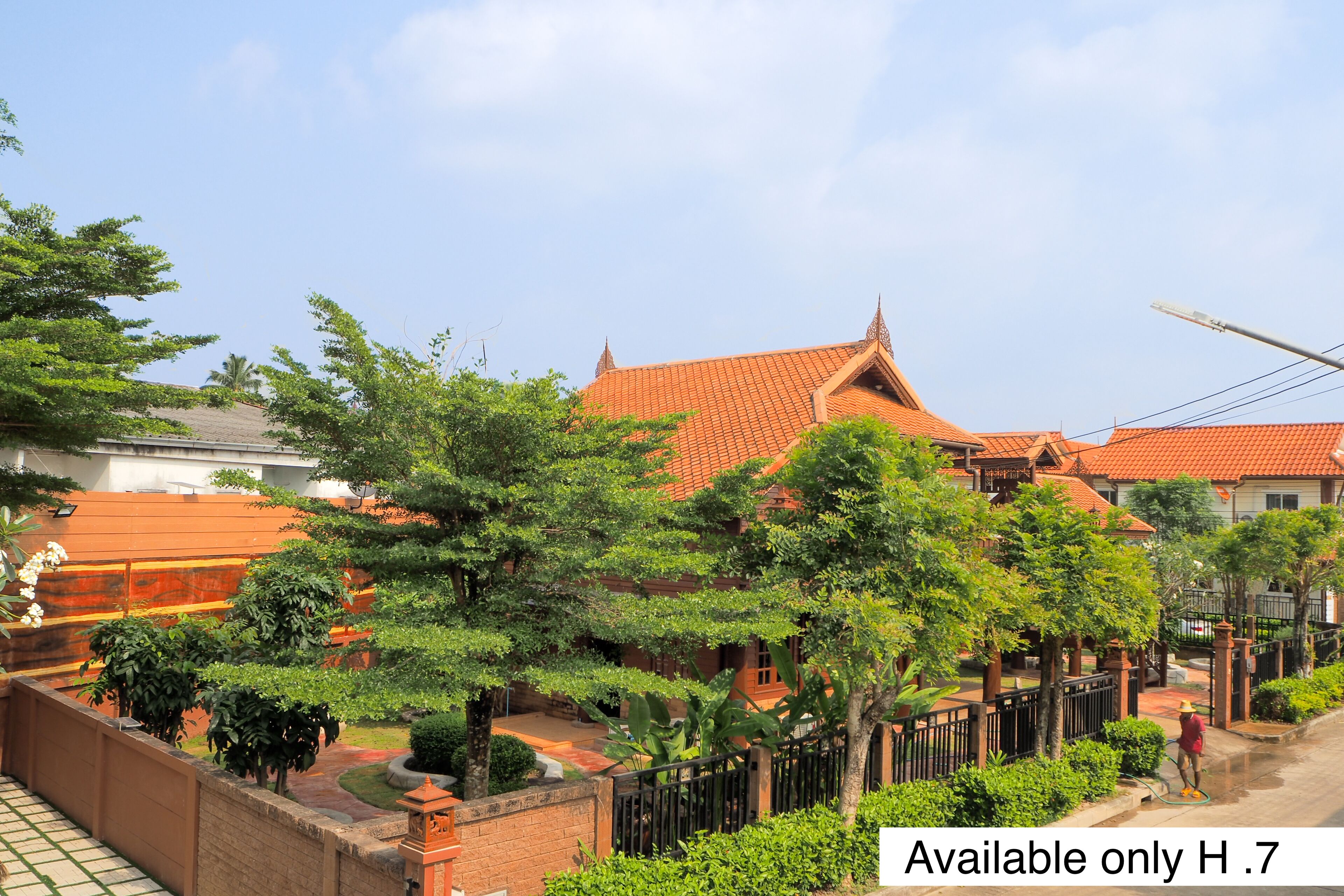 A traditional Thai house with a tiled roof, surrounded by lush greenery and a brick wall.