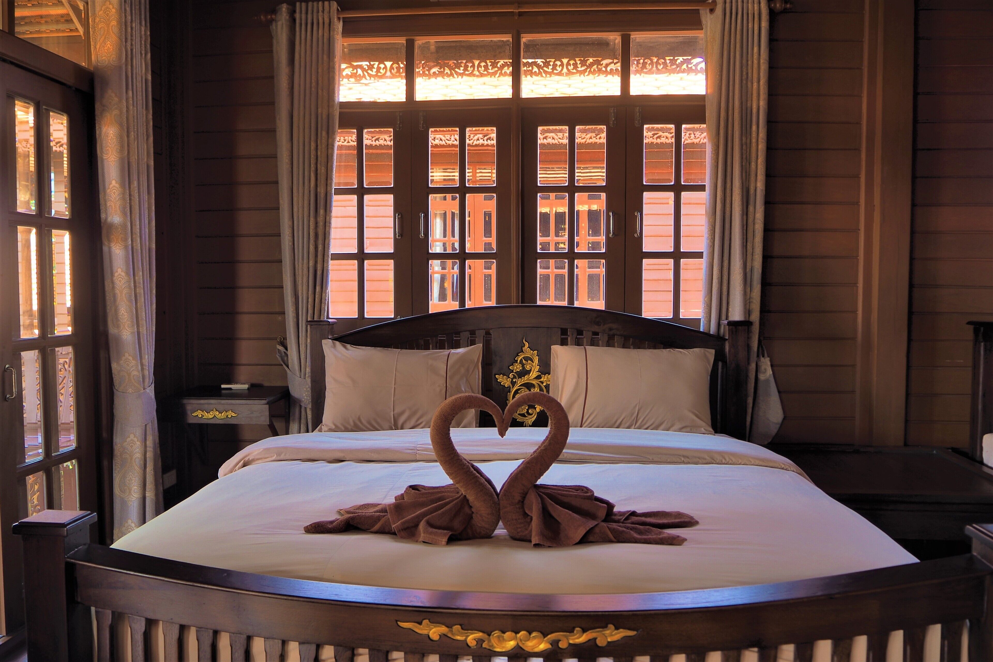 Una cama bien hecha con una disposición de toallas en forma de corazón, un cabecero de madera y una ventana con cortinas decorativas.