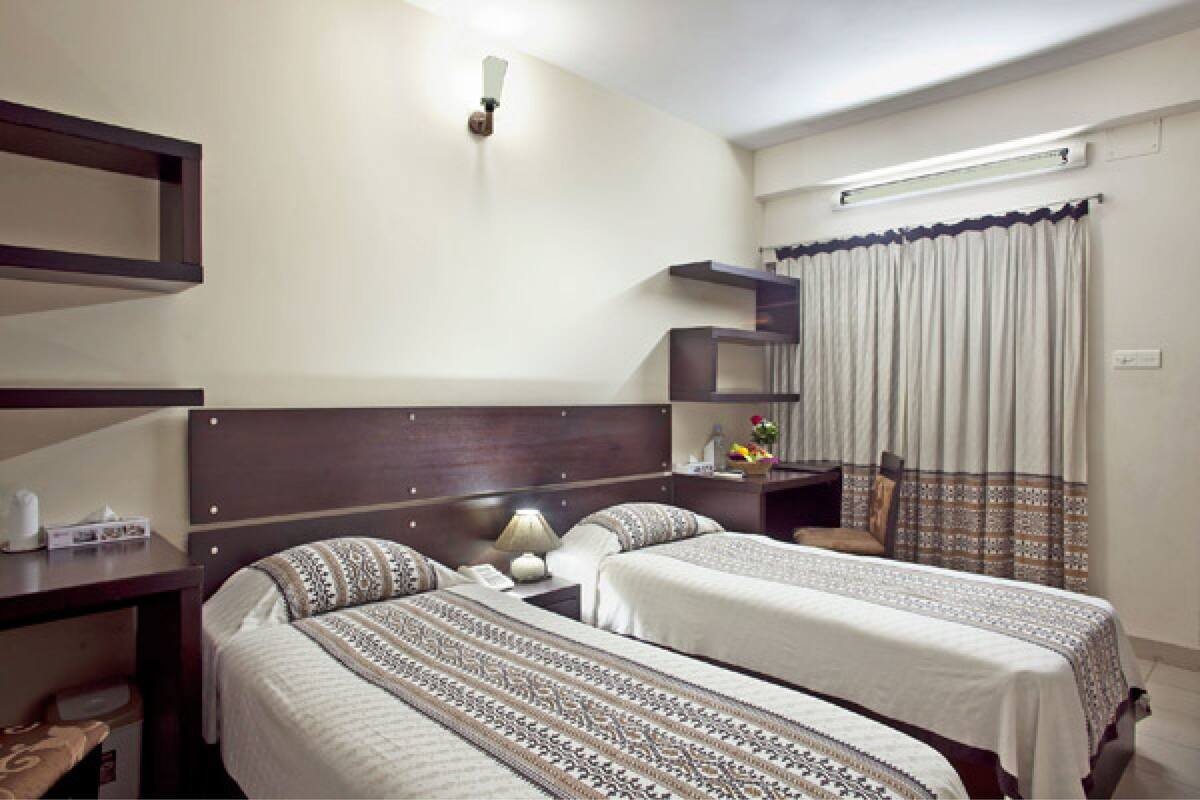 Habitación de hotel con dos camas, una mesita de noche con lámpara, un pequeño escritorio con teléfono y una estantería en la pared.