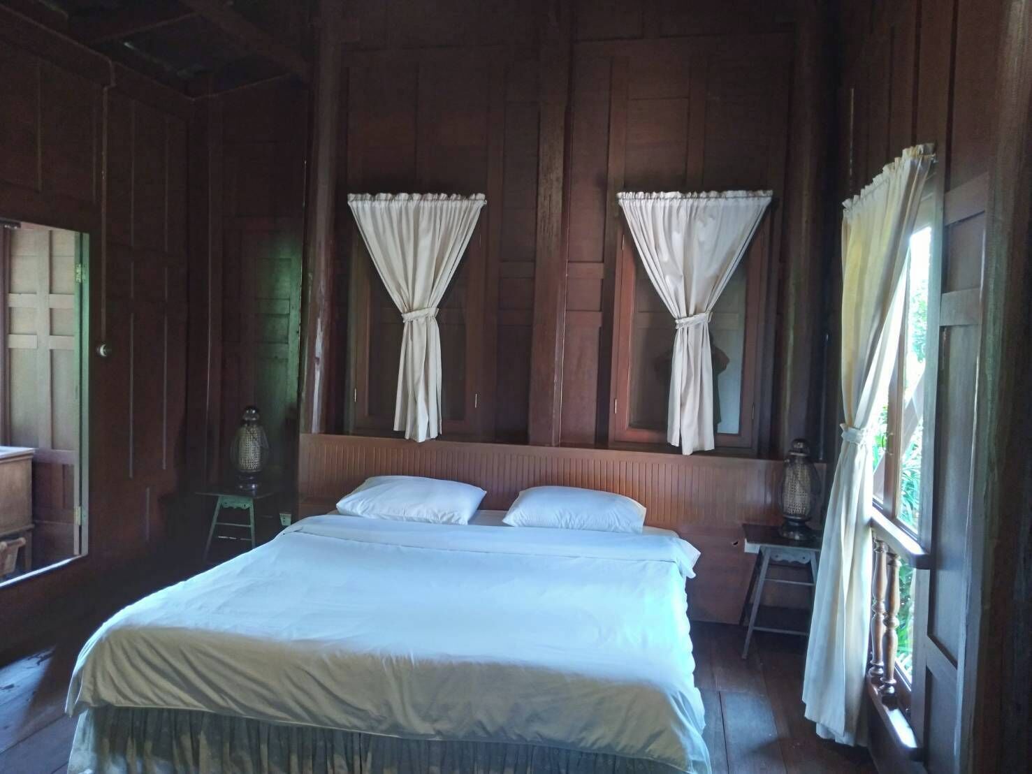 A wooden room with a bed, two windows with curtains, and a nightstand with a lamp.