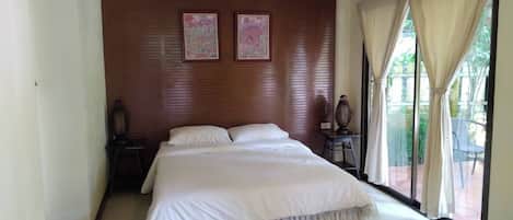 A bedroom with a large bed, a wooden headboard, two wall-mounted pictures, a nightstand with a lamp, and a view of the outdoors through a window with curtains.