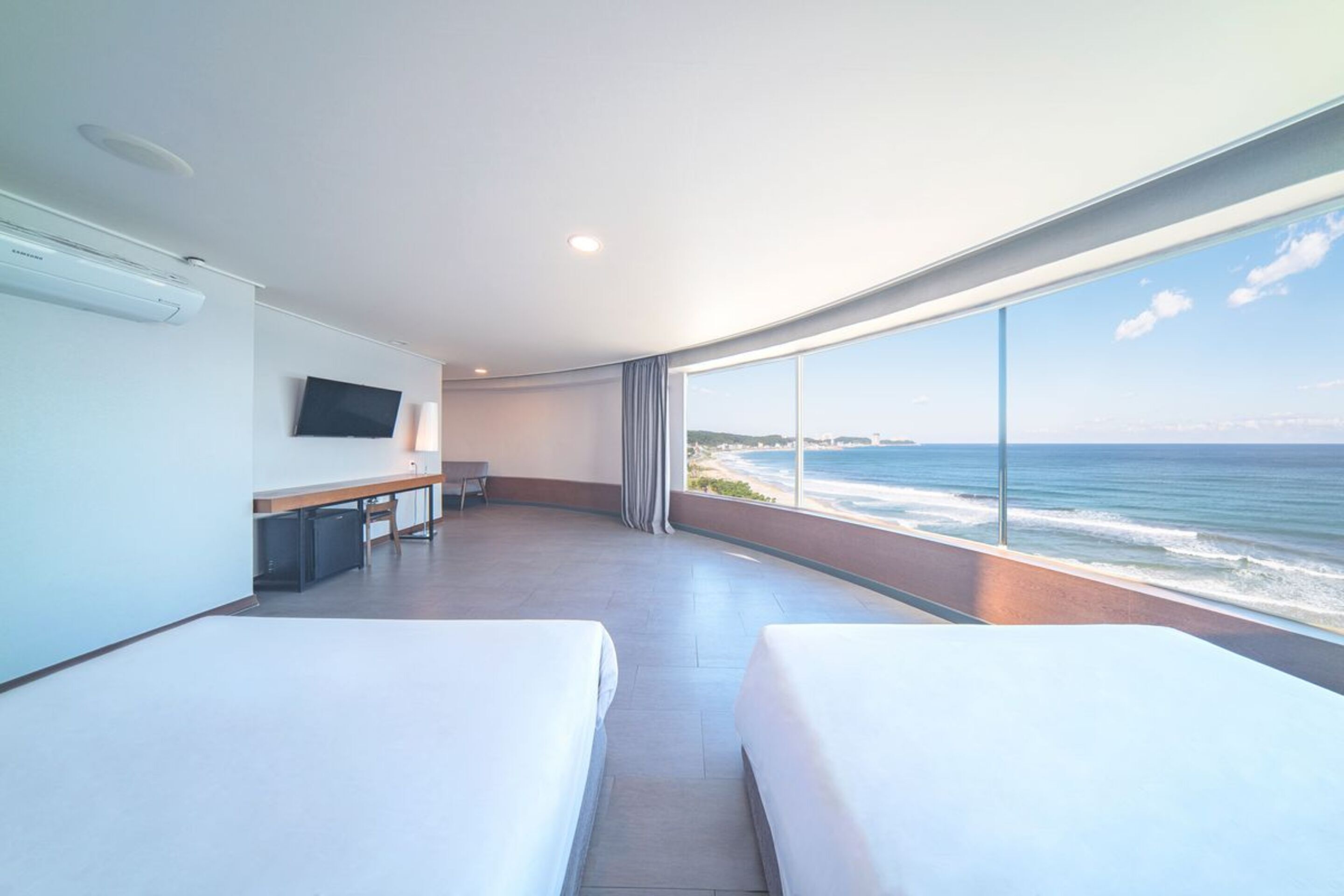 A hotel room with two beds, a desk, a flat-screen TV, and a large window overlooking the ocean.