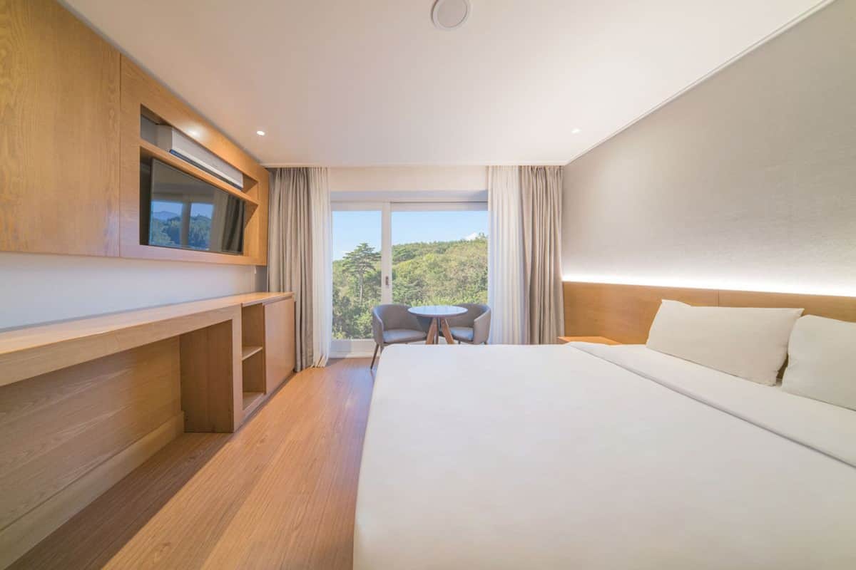 Une chambre d’hôtel avec un grand lit, un bureau en bois, une table ronde et une vue sur la verdure environnante.