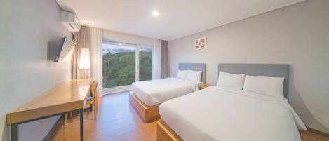 A hotel room with two beds, a desk, and a window with a view of greenery.