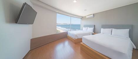 A hotel room with a large window offering a sea view, two beds with white linens, a flat-screen TV mounted on the wall, and a wooden floor.