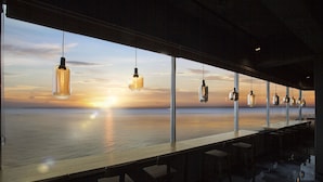 A bar with a view of the ocean at sunset, hanging pendant lights, and a long counter.