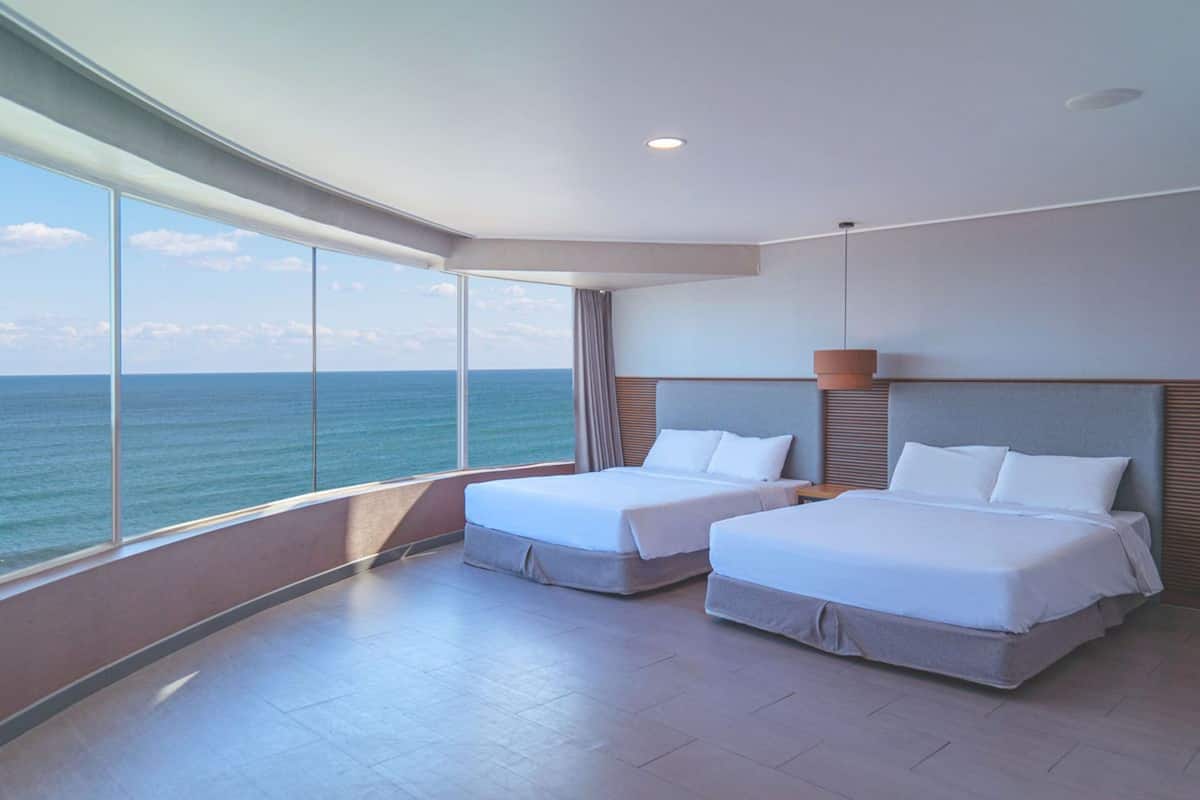 Une chambre d’hôtel moderne avec deux lits, de grandes fenêtres donnant sur l’océan et un plafond incurvé.