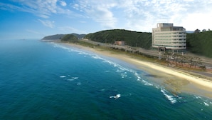 A coastal hotel with a modern design, situated by the beach with a view of the ocean and hills in the background.