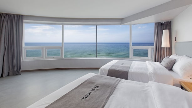 A hotel room with a large bay window, two beds, a bedside table, and a lamp.