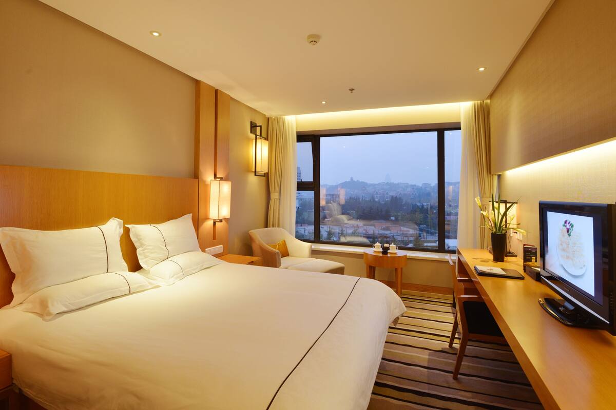 A hotel room with a large bed, a desk with a computer, a chair, and a view of the city through large windows.