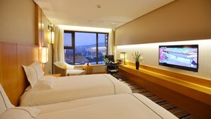 Habitación de hotel con dos camas, un escritorio, una silla, un televisor y un ventanal con vistas a la ciudad.