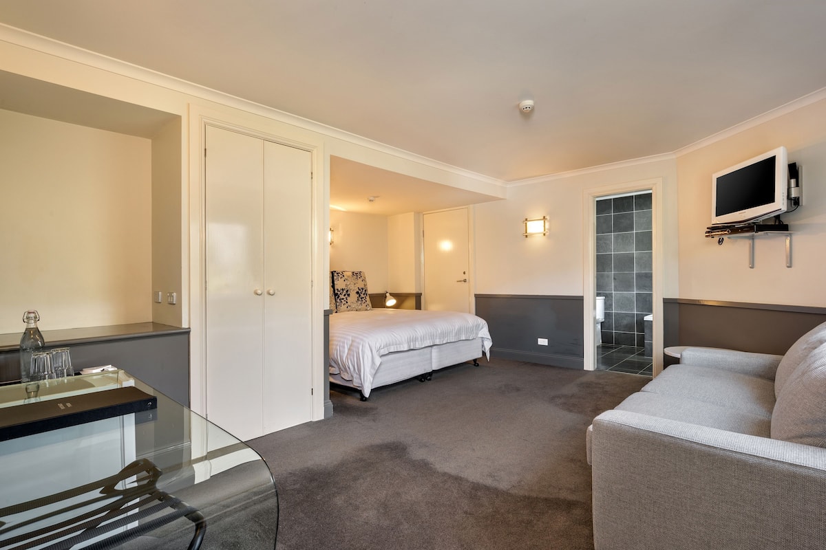 A modern hotel room with a glass dining table, a sofa, a bed, and a TV mounted on the wall.