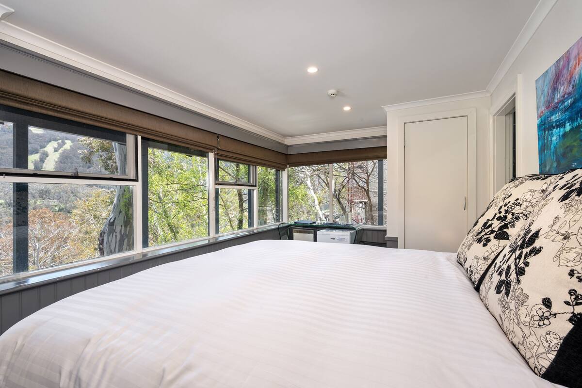 A bedroom with a large bed, a desk, and a view of trees through the windows.