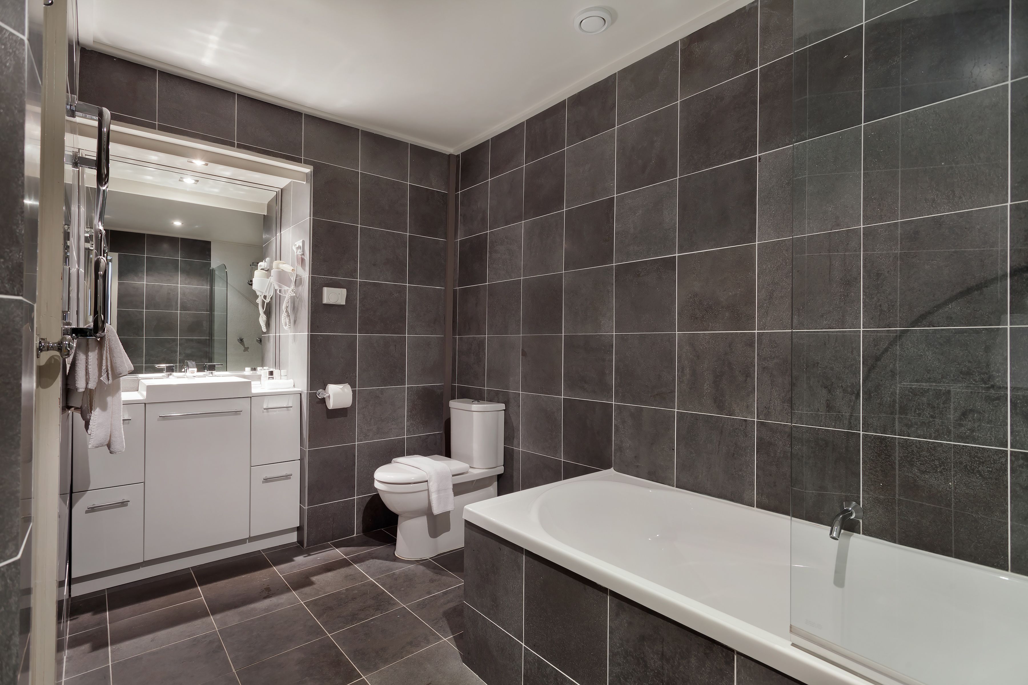 A modern bathroom with dark tiles, a white bathtub, a toilet, and a vanity with a mirror and towel rack.