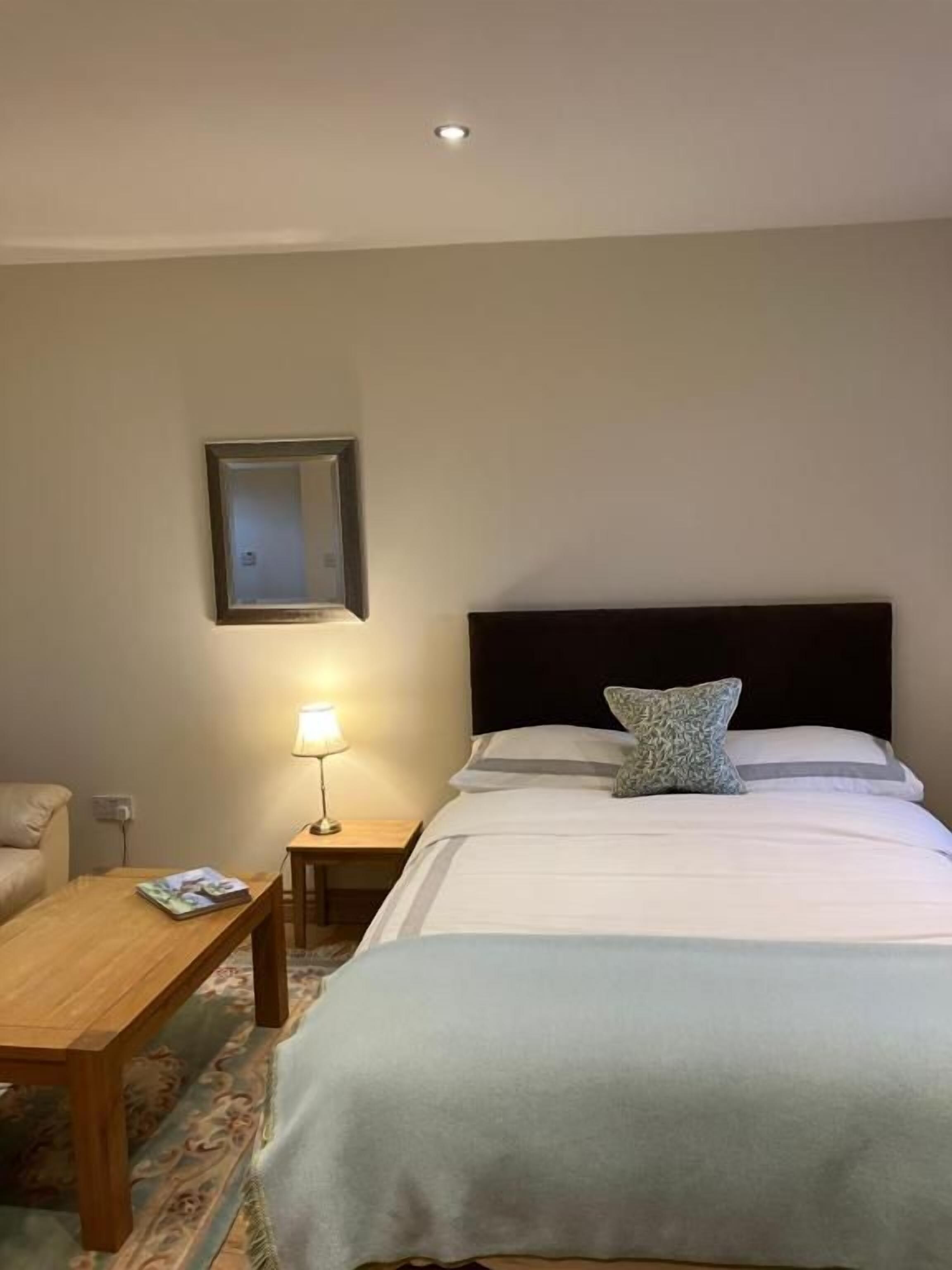 A hotel room with a bed, a nightstand with a lamp, a wooden table, and a framed picture on the wall.