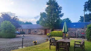 A stone building with a gravel driveway, a wooden bench, a table with an umbrella, and a car parked nearby. - The Courtyard (Perth)