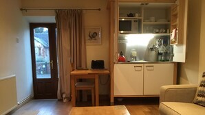 A compact living space with a wooden dining table, a sofa, a kitchenette with a microwave and kettle, and a door leading to an outdoor area. - The Courtyard (Perth)