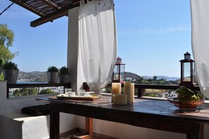 Un balcón con una mesa dispuesta para tomar té, velas y un frutero, con vista a un paisaje costero con plantas en macetas y vistas al mar.