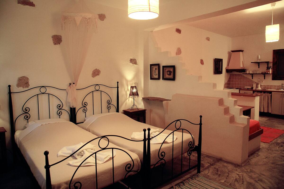 Un dormitorio con una cama doble, una mesita de noche, una lámpara y una pequeña mesa. Se observa una kitchenette al fondo con un lavamanos y una estufa.