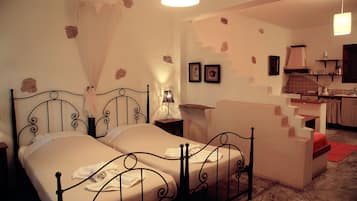 Un dormitorio con una cama doble, una mesita de noche, una lámpara y una pequeña mesa. Se observa una kitchenette al fondo con un lavamanos y una estufa.
