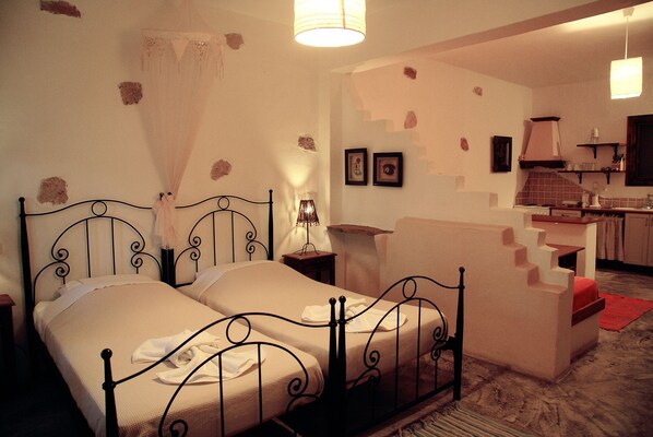 Un dormitorio con una cama doble, una mesita de noche, una lámpara y una pequeña mesa. Se observa una kitchenette al fondo con un lavamanos y una estufa.