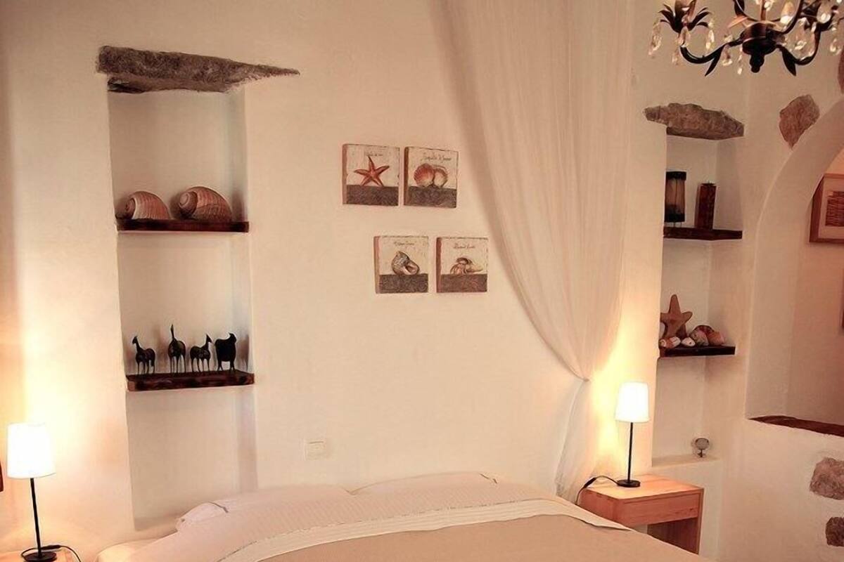 Una cama bien hecha con una colcha color crema y dos toallas dobladas encima. Hay dos mesitas de noche con lámparas, un estante en la pared con objetos decorativos y un candelabro.